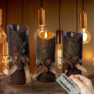 May include: Three brown LED pillar candles with a rustic design, decorated with greenery, acorns, and white ribbon. The candles are lit, and a remote control is in the foreground. The background features hanging light bulbs.
