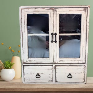 May include: A distressed white cabinet with two glass-paned doors and two drawers. The cabinet has black metal hardware and a rustic, weathered appearance. The cabinet is sitting on a wooden surface.