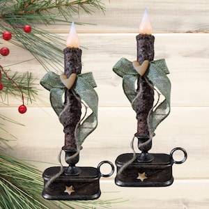 May include: Two rustic-style LED taper candles with a brown, textured appearance. Each candle is adorned with a green burlap bow, a wooden heart, and sits on a black base with a star cutout. The candles have a warm, flickering light.