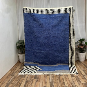 May include: A rectangular, blue rug with a textured surface. The rug features a border of black and white stripes and a design of blue and gray shapes at the bottom. The rug is displayed against a white backdrop.