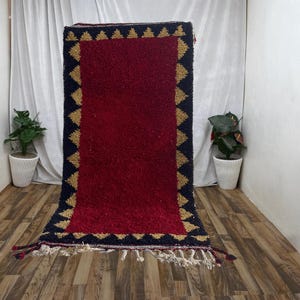 Peut inclure: Un tapis rouge avec une bordure géométrique noire et dorée. Le tapis a des franges sur les bords.