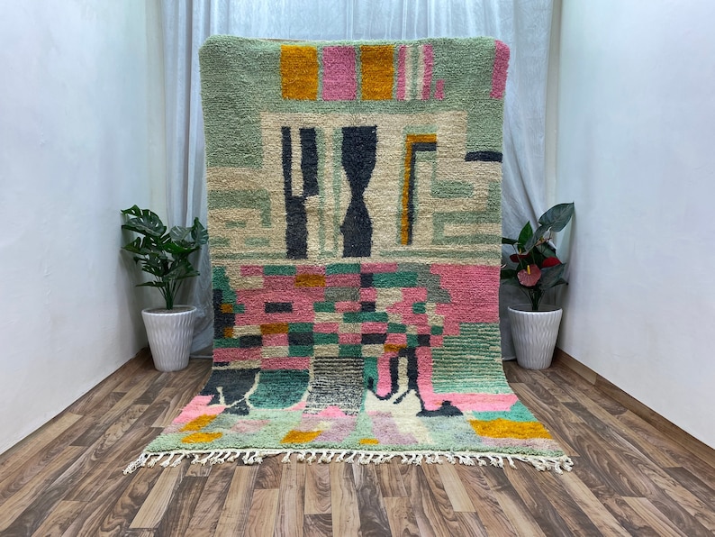 Op de afbeelding: Een kleurrijk abstract tapijt met geometrische vormen in tinten groen, roze, geel en wit. Het tapijt heeft een franje rand en is gemaakt van wol.