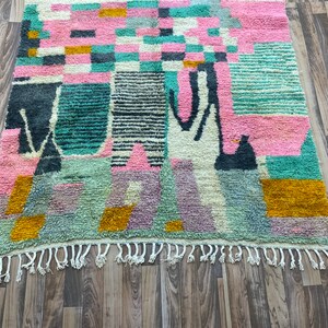 Op de afbeelding: Een kleurrijk abstract tapijt met geometrische vormen in tinten roze, groen, blauw en geel. Het tapijt heeft een franje rand.