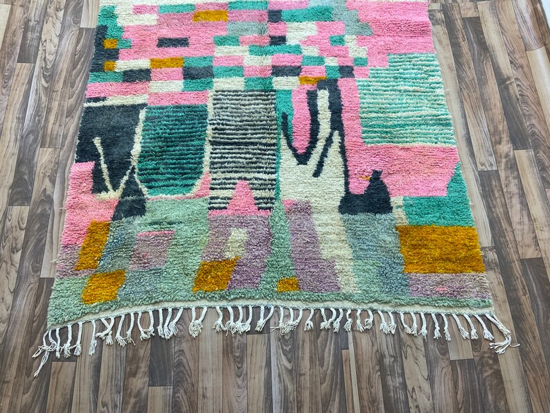Op de afbeelding: Een kleurrijk abstract tapijt met geometrische vormen in tinten roze, groen, blauw en geel. Het tapijt heeft een franje rand.