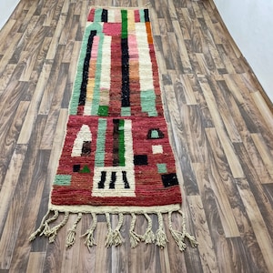 Puede incluir: Una alfombra larga y rectangular con un patrón geométrico en tonos de rojo, verde, negro y blanco. La alfombra tiene flecos en ambos extremos.