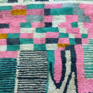Op de afbeelding: Een close-up van een kleurrijk, geometrisch patroon tapijt met roze, groene, zwarte, witte en gouden tinten. Het tapijt is gemaakt van een zacht, pluche materiaal.