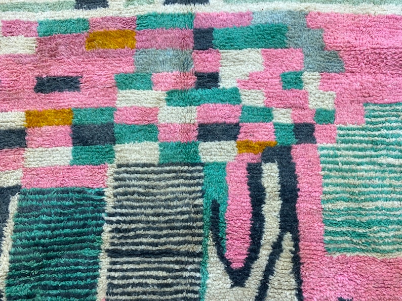 Op de afbeelding: Een close-up van een kleurrijk, geometrisch patroon tapijt met roze, groene, zwarte, witte en gouden tinten. Het tapijt is gemaakt van een zacht, pluche materiaal.