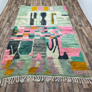 Op de afbeelding: Een kleurrijk abstract tapijt met geometrische vormen in tinten roze, groen, geel en zwart. Het tapijt heeft een franje rand.