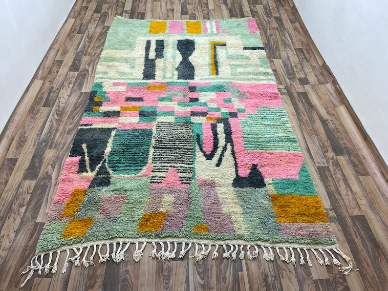 Op de afbeelding: Een kleurrijk abstract tapijt met geometrische vormen in tinten roze, groen, geel en zwart. Het tapijt heeft een franje rand.