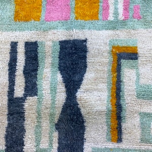 Op de afbeelding: Close-up van een kleurrijk tapijt met geometrische patronen in tinten blauw, groen, geel en roze. Het tapijt heeft een textuur, een pluche oppervlak.