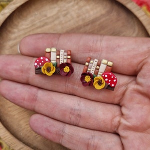 Peut inclure: Une paire de boucles d'oreilles en argile polymère faites à la main avec une pile de livres, un champignon et des fleurs dans des tons de rouge, jaune et marron.