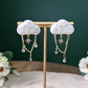 May include: A pair of white cloud-shaped earrings with gold chains and star charms. The chains dangle from the bottom of the clouds and feature small crystal accents.