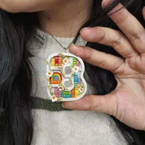 May include: A white ceramic necklace pendant shaped like the letter "B". The inside of the pendant is decorated with a rainbow, flowers, and colourful books on a bookshelf.