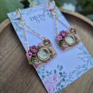 May include: Handmade earrings in the shape of cameras, with tan bodies, gold glitter lenses, and purple and pink floral embellishments. The earrings are suspended from gold-tone chains and hooks. The backing card displays the text "DIVINE By Ky".