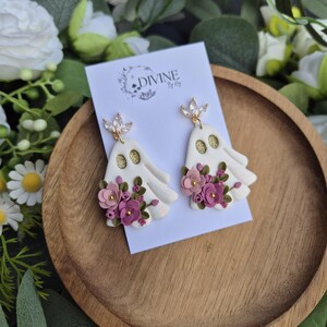 Op de afbeelding: Witte, spookvormige oorbellen met gouden glitteraccenten en bloemversieringen. De oorbellen hebben een gouden kroonachtige top en worden gepresenteerd op een witte kaart met de tekst "DIVINE By Klg".