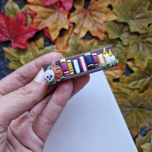 Op de afbeelding: Een miniatuur boekenkast met een bruine basis, versierd met een kleine oranje pompoen, een witte geest en kleurrijke boeken. De boekenkast is versierd met kleine witte kaarsen en groene bladeren.