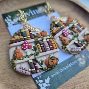 May include: A pair of round dangle earrings featuring a miniature bookshelf design with colourful books, orange pumpkins, and a red and white spotted mushroom. The earrings are crafted with gold-toned metal hooks and green vine accents on a white background.