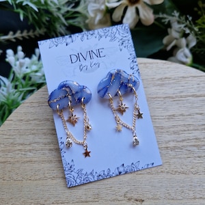 Puede incluir: Un par de pendientes de nube azul y oro con cadenas colgantes y estrellas. Los pendientes están en una tarjeta blanca con el texto "DIVINE by Kay".