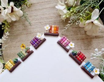 May include: A set of six miniature bookshelf decorations with different colored books and flowers. Each bookshelf is made of brown clay and features a small plant or flower.
