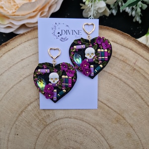 Könnte beinhalten: Ein Paar schwarze herzförmige Ohrringe mit einem Totenkopf, Blumen und Büchern. Die Ohrringe sind aus Polymer Clay gefertigt und haben goldene Akzente. Die Ohrringe sind mit "DIVINE by" beschriftet.