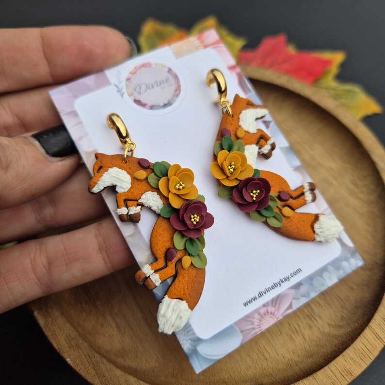 May include: A pair of fox-shaped dangle earrings featuring orange clay foxes with white-tipped tails and chests, adorned with small yellow, burgundy, and green clay flowers. The earrings have gold-tone metal hooks and are displayed on a white card.