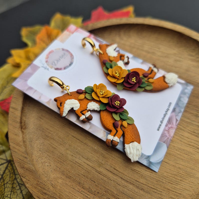 May include: A pair of orange fox earrings with white accents, adorned with yellow, burgundy, and green floral details. The earrings have gold-colored hooks and are displayed on a card with the text "Divine" and a website address.