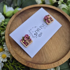 Op de afbeelding: Een paar oorbellen van polymer clay met een stapel boeken met roze bloemen erop. De oorbellen worden getoond op een witte kaart met de tekst "DIVINE By Kr" erop gedrukt.