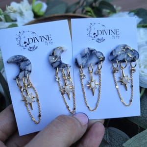 May include: A pair of earrings featuring a crescent moon and a cloud, both with a marbled black and white design and gold accents. Each earring has a dangling star charm with a white stone and a gold chain with small gold beads.