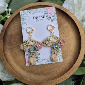 May include: A pair of whimsical mushroom earrings with floral accents. The earrings feature a beige cap with gold glitter, adorned with pink and blue flowers. The earrings are displayed on a card with the text "DIVINE By Kay".