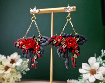 Könnte beinhalten: Ein Paar schwarze Hängeohrringe mit roten und goldenen floralen Akzenten. Die Ohrringe haben eine zarte Kette und einen kleinen Kristall oben.