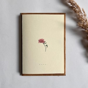 May include: A cream-coloured greeting card with a brown border, featuring a small pink flower illustration. The word "Love" is printed at the bottom. A dried plant stem is positioned to the right of the card.