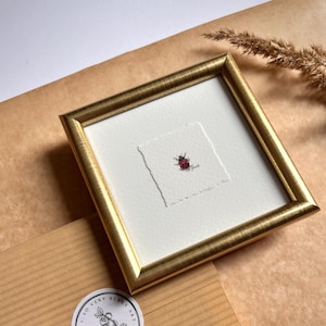 May include: A framed miniature watercolor painting of a red ladybug with black spots. The painting is set in a gold-colored frame on a white background. A brown paper envelope with a round sticker that says "So Very Berry Art" is in the foreground.