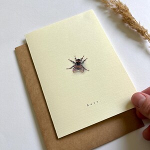 Può includere: Biglietto di auguri color crema con un'illustrazione dettagliata di un'ape. La parola "Buzz" è stampata sotto. Il biglietto è appoggiato su una busta marrone, con un filo d'erba secca sullo sfondo.