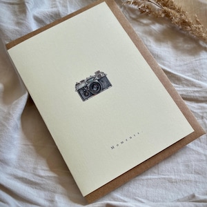 Peut inclure: Une carte de vœux rectangulaire blanc cassé présentant une petite illustration détaillée en noir et blanc d'un appareil photo vintage centrée dans la partie supérieure. Sous l'appareil, le mot "Moments." est imprimé en police noire.