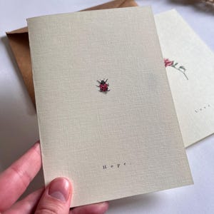 Puede incluir: Dos tarjetas de felicitación color crema. Una tarjeta presenta una pequeña ilustración de mariquita roja y la palabra "Hope". La segunda tarjeta tiene un diseño floral. Un sobre marrón es visible detrás de las tarjetas. Las tarjetas tienen una superficie texturizada.