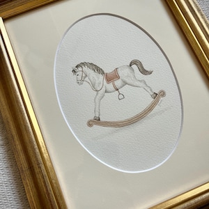 Könnte beinhalten: Ein weißes Schaukelpferd mit braunem Sattel und braunem Sockel, in Aquarell gemalt und in einem goldenen Rahmen gerahmt.