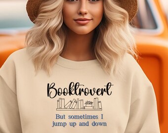 May include: A cream-colored sweatshirt with the text "Booktrovert" above a row of book illustrations. Below, the text reads "But sometimes I jump up and down." The sweatshirt is worn by a person in front of an orange vintage truck.
