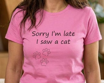 May include: A pink short-sleeved t-shirt featuring the black text Sorry I'm late I saw a cat and a graphic of three small cat paw prints. The shirt has a crew neckline and is paired with blue denim jeans, creating a casual everyday look.