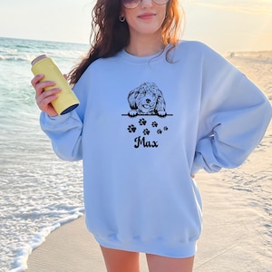 May include: Light blue sweatshirt with a black and white illustration of a dog peeking over the top. The dog is surrounded by paw prints and the name "Max" is printed below.