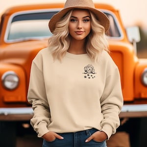 May include: A woman wearing a beige sweatshirt with a small embroidered image of a dog peeking out of a pocket. The dog is brown and white and has paw prints embroidered below it.