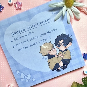 May include: Square sticky notes with a light blue heart-shaped background and a cartoon illustration of two figures. The text reads "Square sticky notes", "Sticks well!!", and "Doesn't leave glue marks on the note under !!".