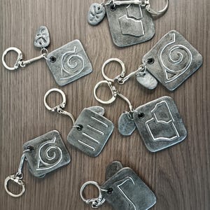 Peut inclure: Ensemble de dix porte-clés argentés avec des motifs différents. Chaque porte-clés est doté d'un charme de forme carrée avec un symbole différent, notamment une spirale, une feuille et une main. Les charmes sont attachés à des anneaux de clés argentés.