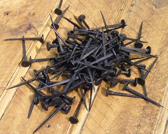 May include: A pile of black metal nails with pointed tips and flat heads. The nails are scattered on a wooden surface.