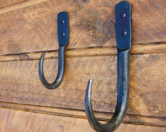 May include: Two black metal hooks mounted on a wooden wall. The hooks are curved and have a rustic, hand-forged look.