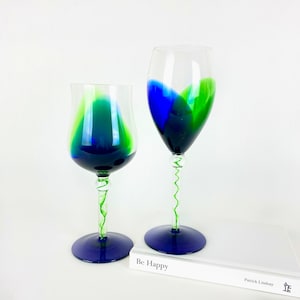 May include: Two glass wine glasses with blue and green swirls on the stem and bowl. The glasses are sitting on a white book with the text "Be Happy" on the cover.