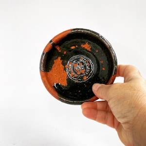May include: A small, round, terracotta dish with a black glaze. The dish has a rough, textured surface and a slightly raised rim.