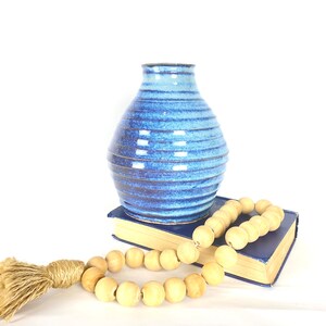 May include: A blue ceramic vase with a textured, striped design. The vase is sitting on a blue book with a wooden bead garland draped over it.