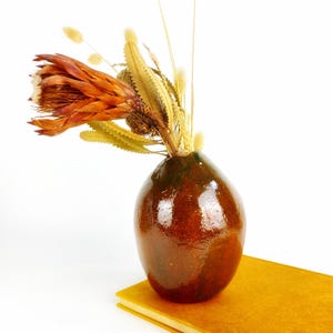 May include: A brown, glossy ceramic vase with dried flowers. The vase is rounded and rests on a yellow book. The dried flowers include an orange protea and beige and green stems. The vase is approximately 15 cm tall.