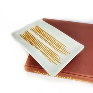 May include: A white rectangular ceramic dish with a gold stripe design. The dish is sitting on top of a brown book.