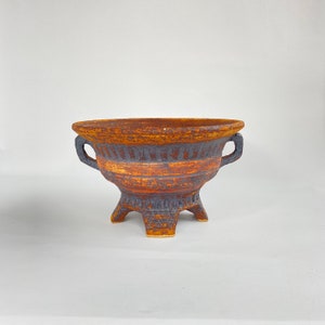 May include: A brown and blue ceramic planter with three legs and two handles. The planter has a textured surface and a striped design.